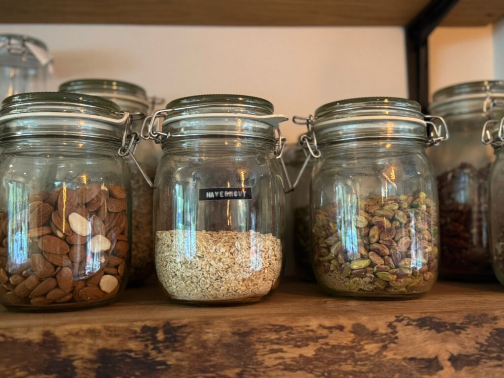 Een rij glazen potten met amandelen, havermout en pistachenoten op een houten plank.